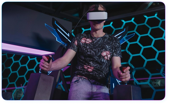 Młody mężczyzna w goglach wirtualnej rzeczywistości steruje rozgrywką za pomocą dwóch joysticków, siedząc w fotelu symulatora na tle ściany z neonowymi plastrami.