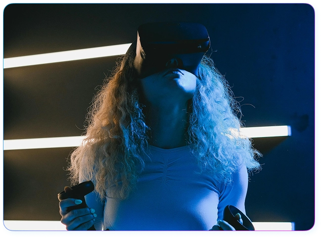 Kręconowłosa dziewczyna w goglach VR i z joystickami w rękach spogląda w górę na tle poziomych neonów.